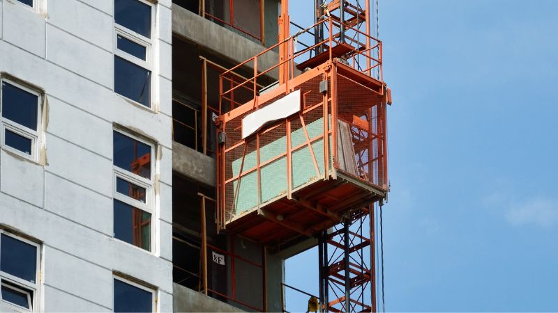 Un ascensor de construcción de color naranja asciende por un edificio de gran altura en construcción, con ventanas visibles y exterior de hormigón.