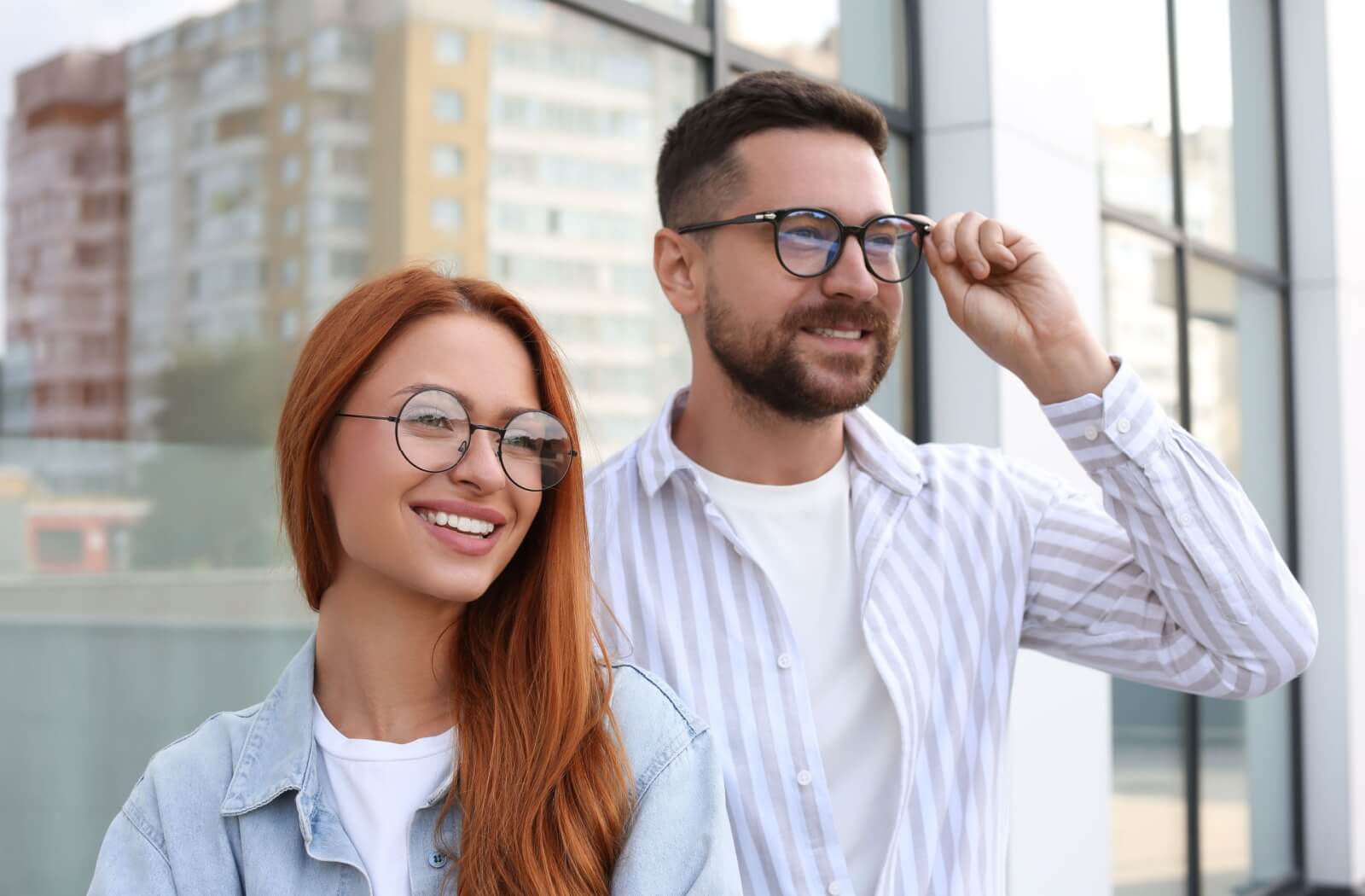 A well-dressed couple, one in round eyeglasses and the other in black-rimmed glasses, smile out at a city landscape