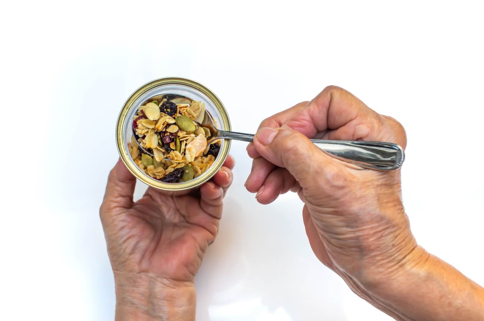 A close-up top view of an older adult's right hand scooping out a spoonful of nuts and seeds from a bowl in their left hand.