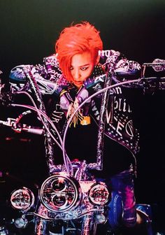 This  contain an inage of   G-Dragon with red hair sitting on top of a motor bike in front of a black background