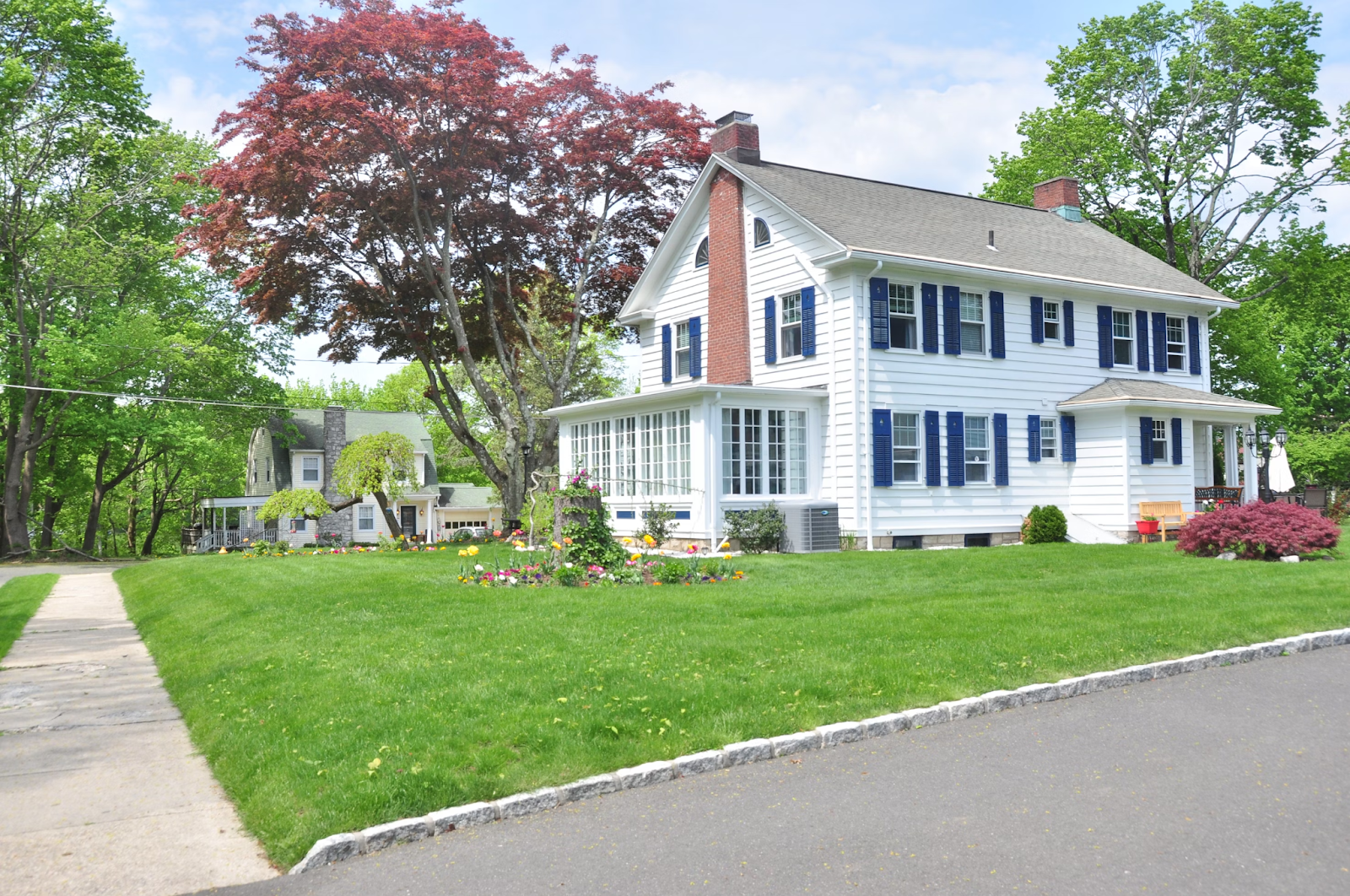 A house with white vinyl siding, a brick chimney and blue shutters is pictured during the daytime. A green lawn surrounds the home.