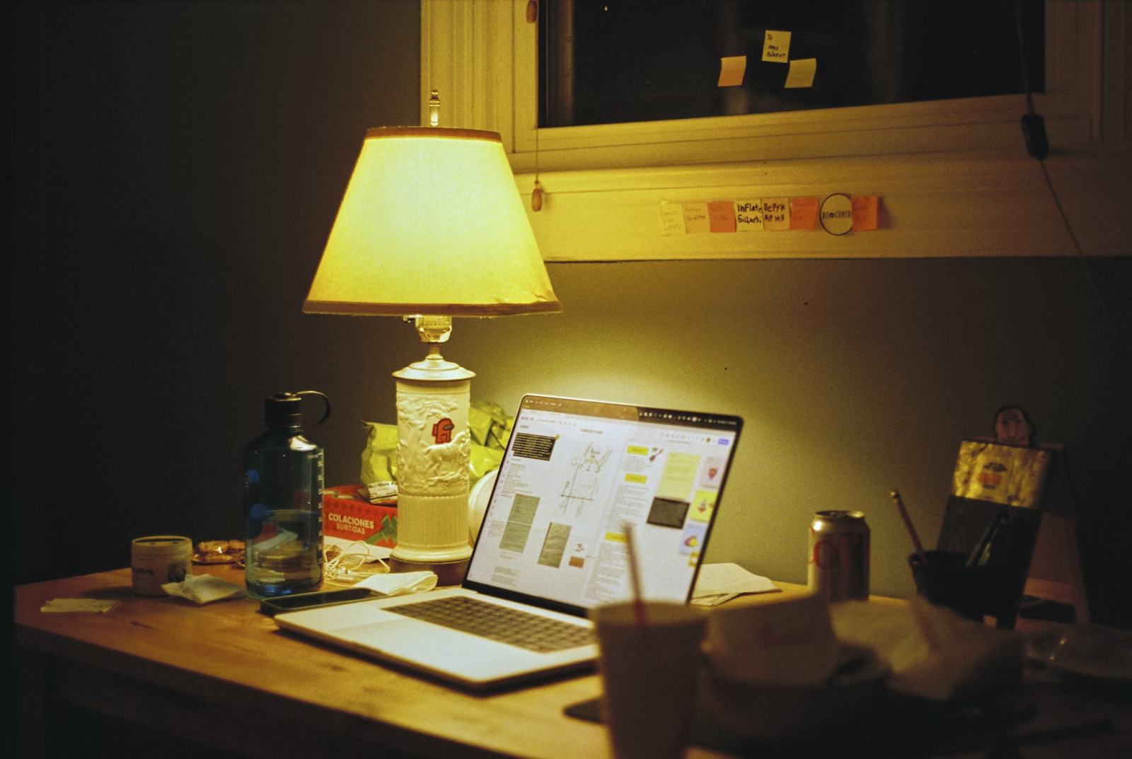 An open laptop sits on a desk with a lamp lighting up the surrounding area. A takeaway box and drink sit to the side, showing that someone was hard at work on writing essays that impress.