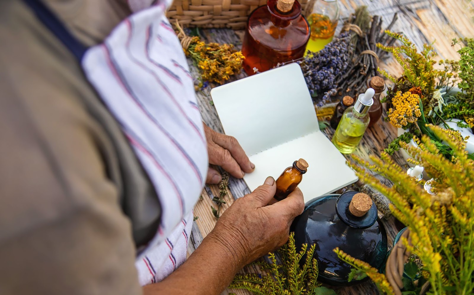 An unrecognized herbalist carefully prepares tinctures from medicinal herbs, holding a notebook and an empty amber glass bottle.