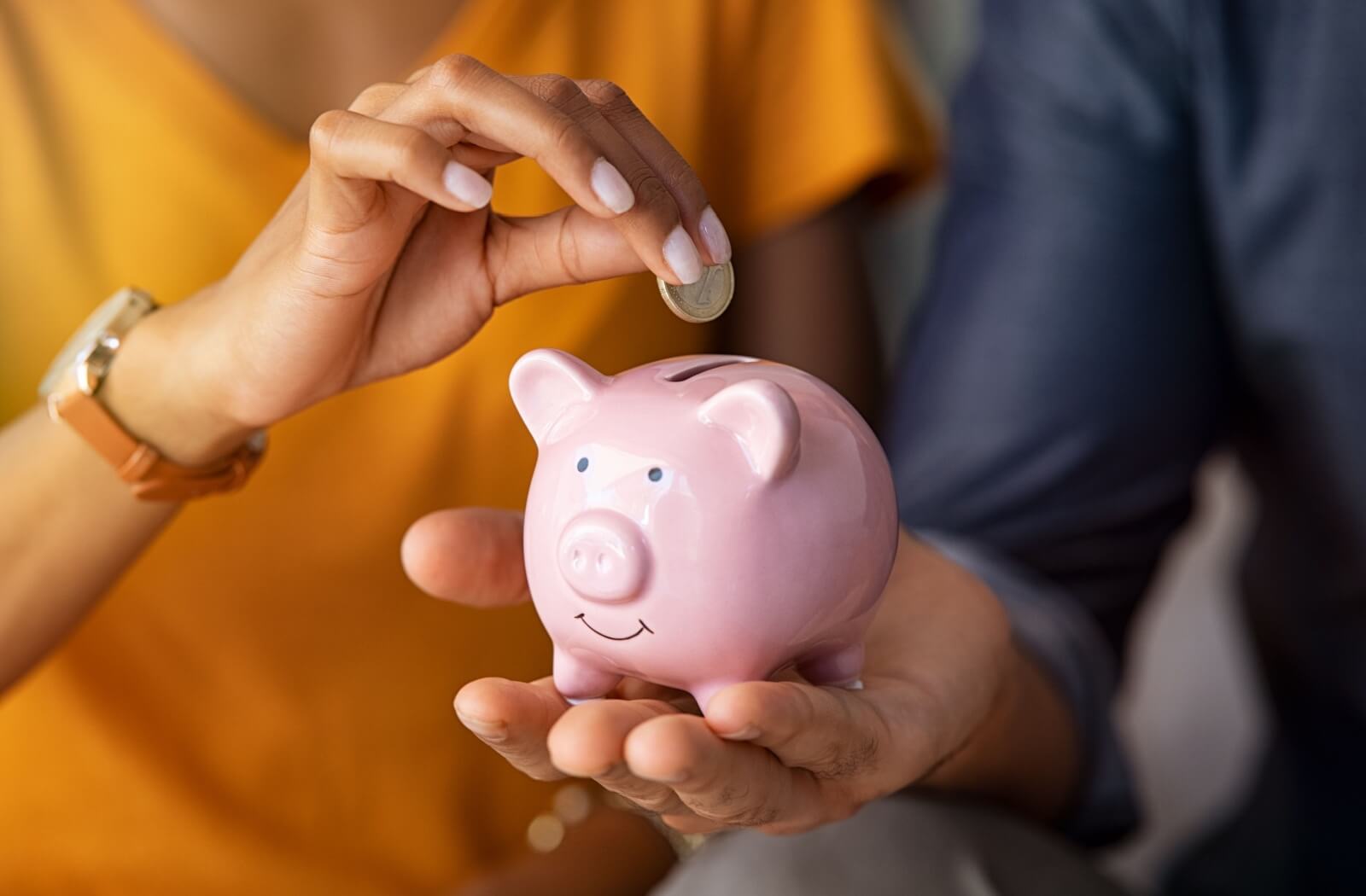 A couple blurred in the background holding an in-focus piggy bank while putting money in it.