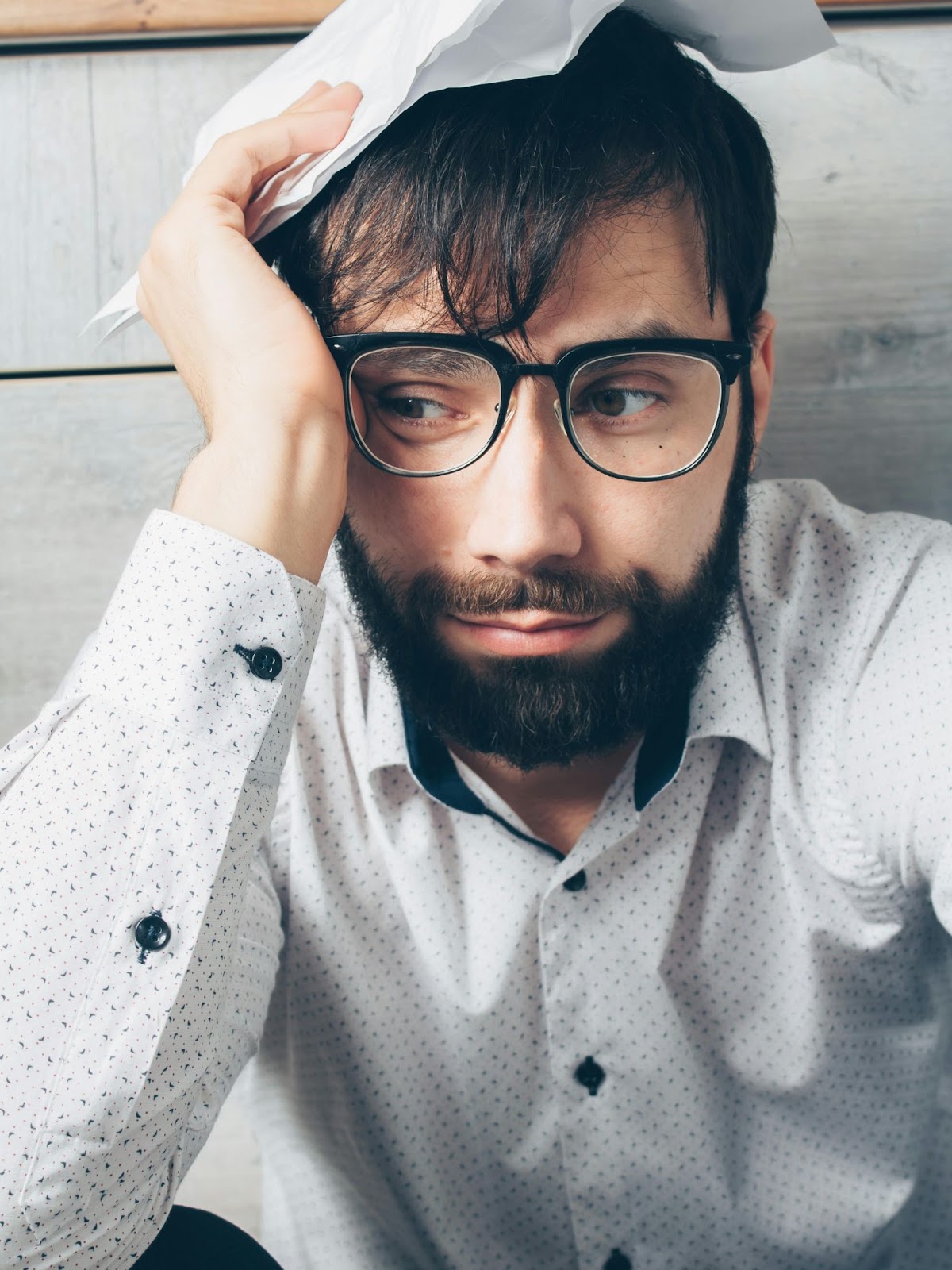 Image shows a bearded white man, with dark hair, wearing glasses and a white shirt with black buttons. He is resting his head on one hand in a gesture of despair.