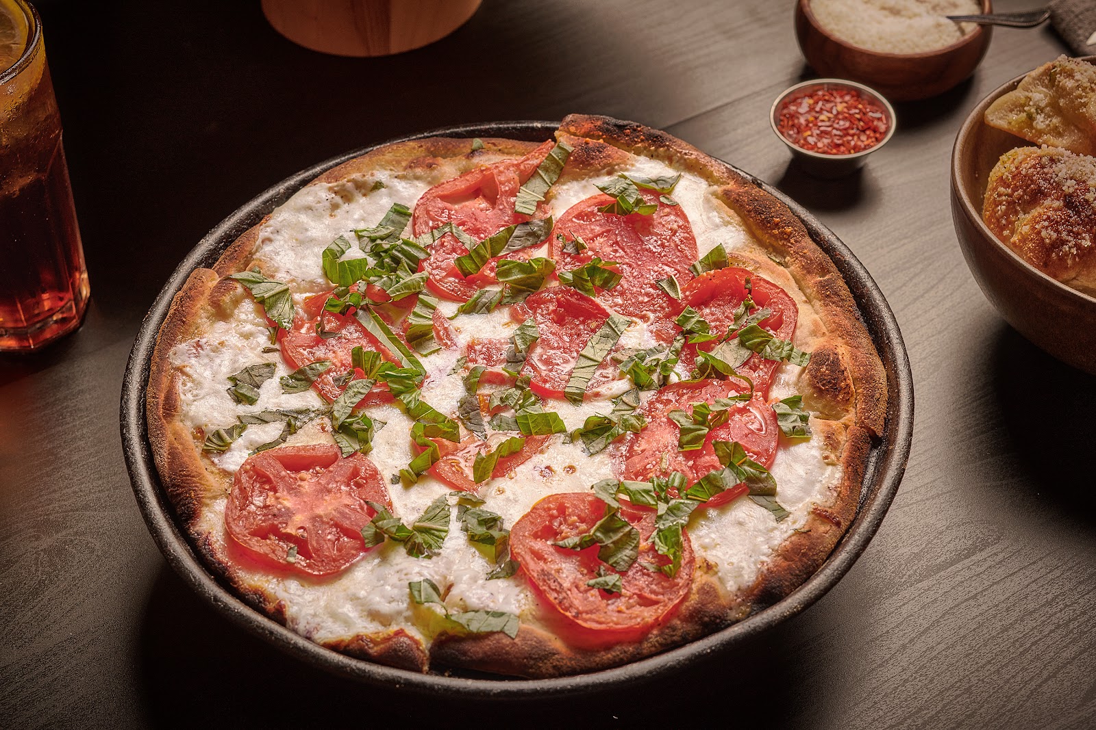 A classic Margherita pizza with fresh mozzarella, sliced tomatoes, and basil on a crispy crust, served with garlic knots and red pepper flakes.