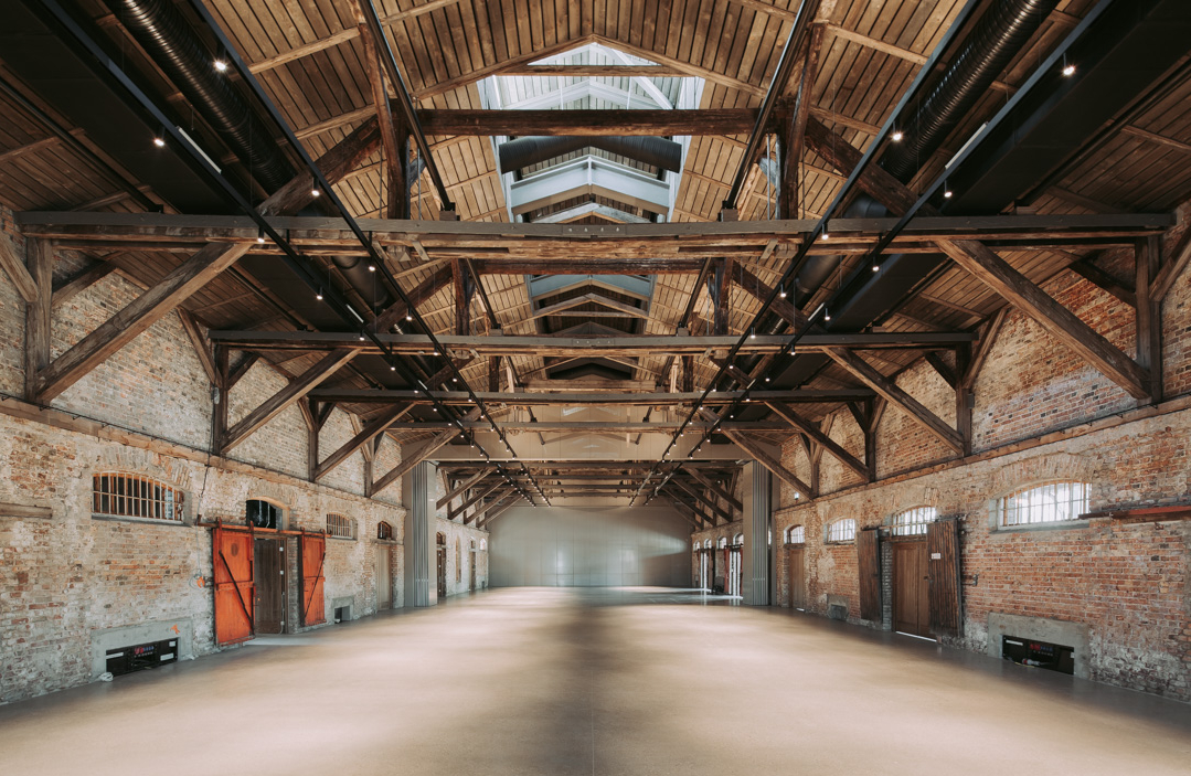 A spacious industrial-style interior with exposed brick walls and a high wooden beam ceiling showcases the artistry of industrial adaptive reuse.