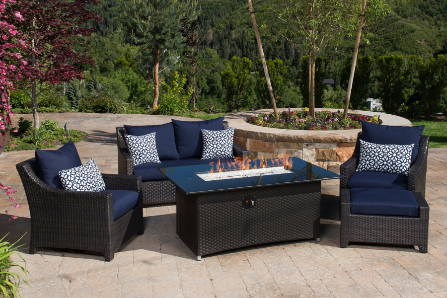 A sleek outdoor dining set with a large rectangular table and eight wicker chairs featuring navy blue cushions, arranged on a sunlit patio overlooking a pool and scenic hillside views.