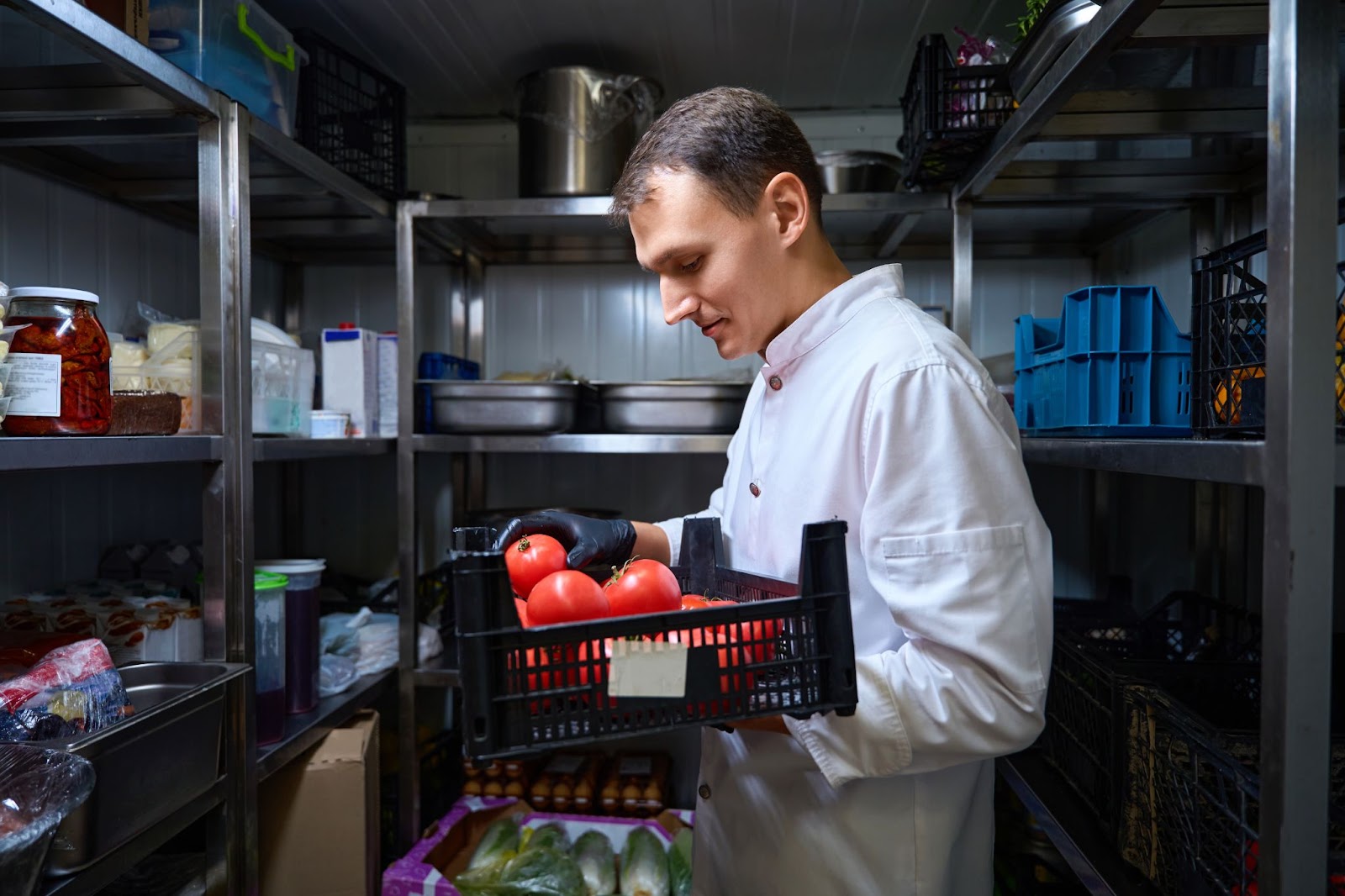 Kitchen worker looking down at a crate of tomatoes in his hands in a walk-in commercial refrigerator.