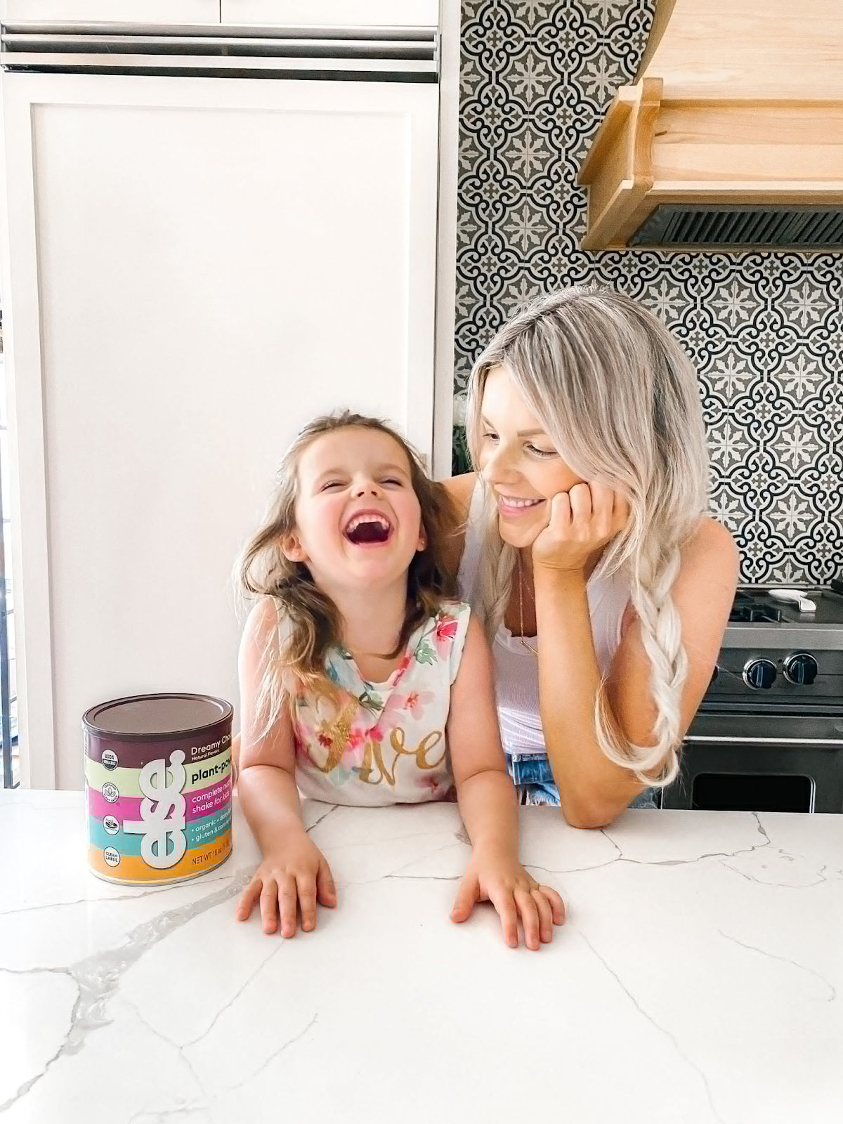 A woman and a little girl are joyfully laughing together in a cheerful kitchen, surrounded by homey decor.