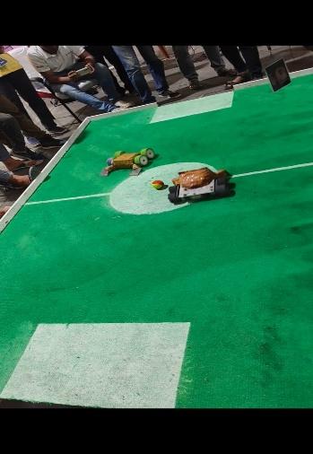 Two student-built robots competing in a robotic soccer match on a green playing field surrounded by onlookers.