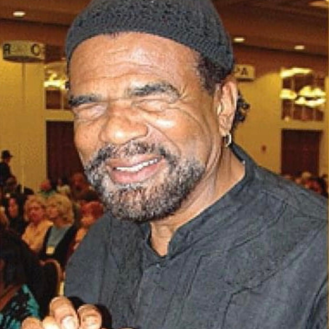 A black man stands in a conference hall, smiling at the camera. He has a short black goatee peppered with white hair. He wears a grey knit beanie and a grey shirt