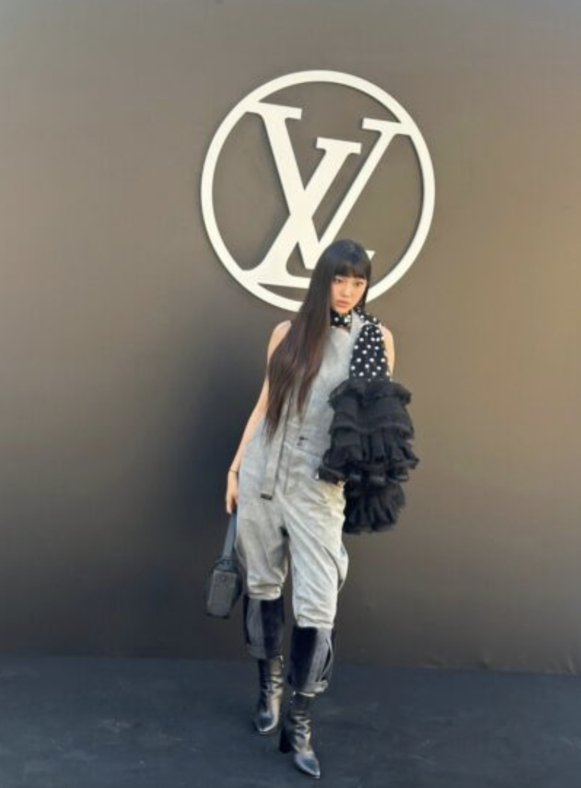 A picture of NewJeans' Hyein wearing ash color jumpsuit, black boots and a black handbag  