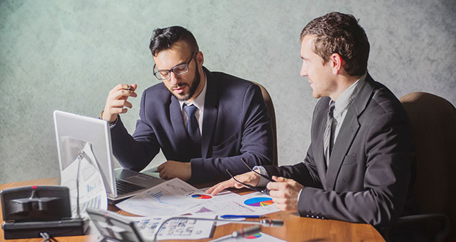 Image contains two writing professionals in black suits, discussing a topic with papers on the table.