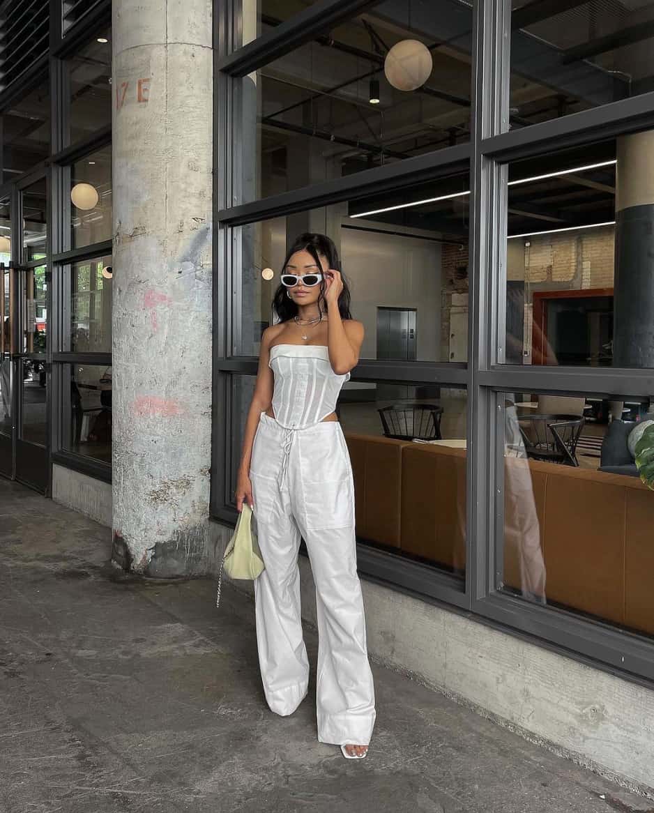 A woman wearing white cargo pants, a white corset top, and white heels and sunglasses with a light yellow handbag