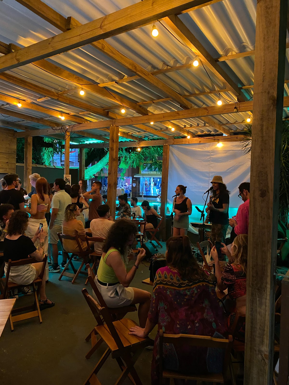 Vários grupos de pessoas sentados em mesas, reunidos no Lobitos Bar, curtindo um show ao vivo. 