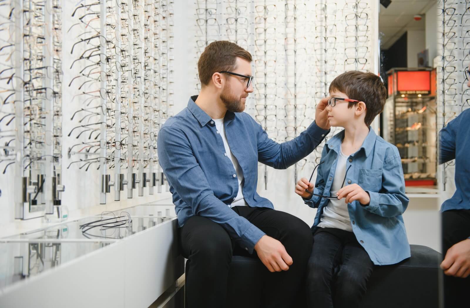 A parent sits with their child at an optometrist’s to help their child try on new eyeglasses after an eye appointment.