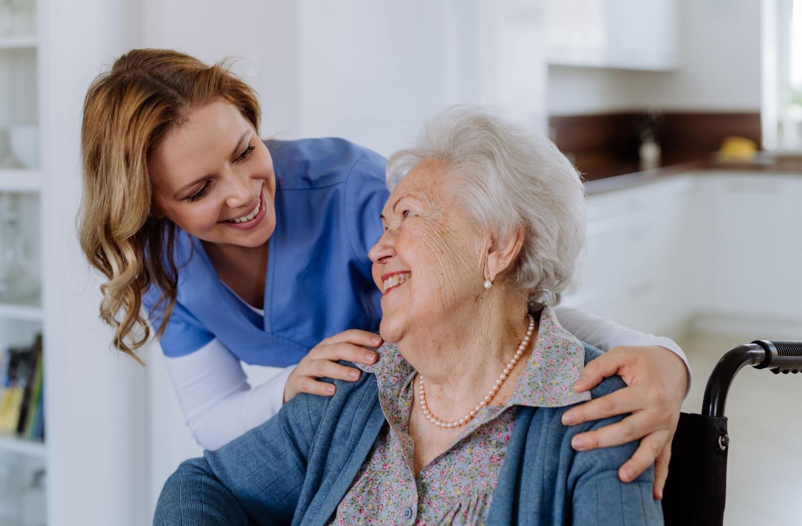 A smiling caregiver interacts warmly with an elderly woman in a wheelchair.