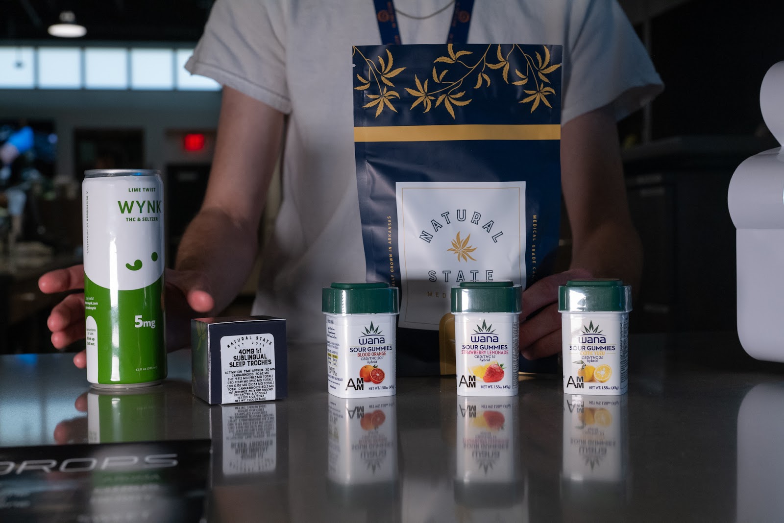 Several cannabis products appear close up in the frame, including a Wynk cannabis infused beverage, several boxes of Wana gummies with high CBD content and a tincture by Natural State Medicinals.