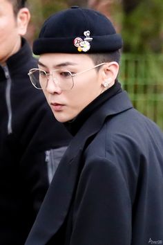 This contains an image of G-Dragon wearing glasses and a black hat is seen in front of two other men