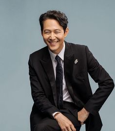 This contain Lee Jung Jae in a suit and tie sitting on a stool smiling at the camera with his hands behind his back