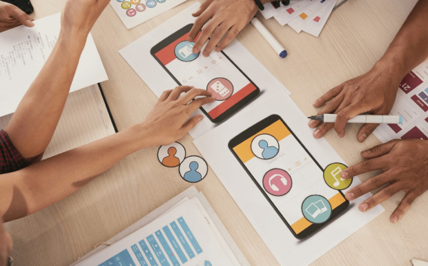 A team of people collaborating on a mobile app design. They are gathered around a table, reviewing sketches and discussing ideas about Mobile App Development Process