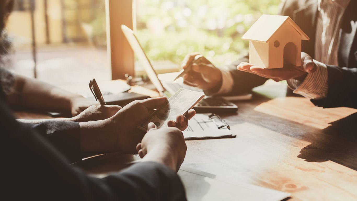 A close-up of individuals holding a small model house and exchanging money across a table during a real estate transaction.