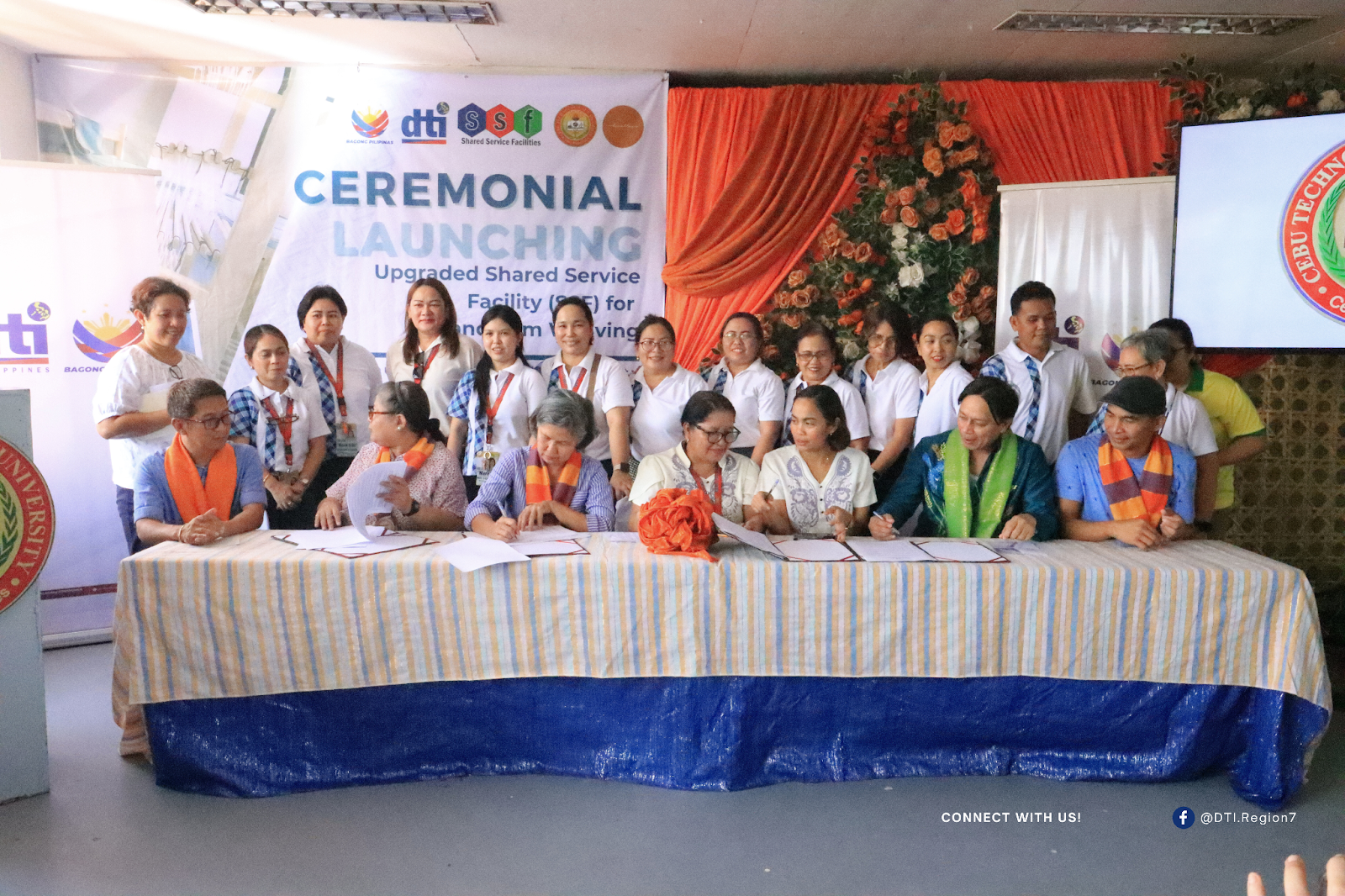 A group of people participating in the ceremonial launching of the Upgraded Shared Service Facility for Food Processing and Weaving, with a banner in the background and documents being signed at a table.