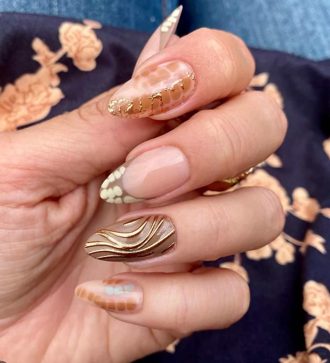 Close-up of a hand with detailed nail art, featuring gold accents and patterns on a blue floral fabric background.

