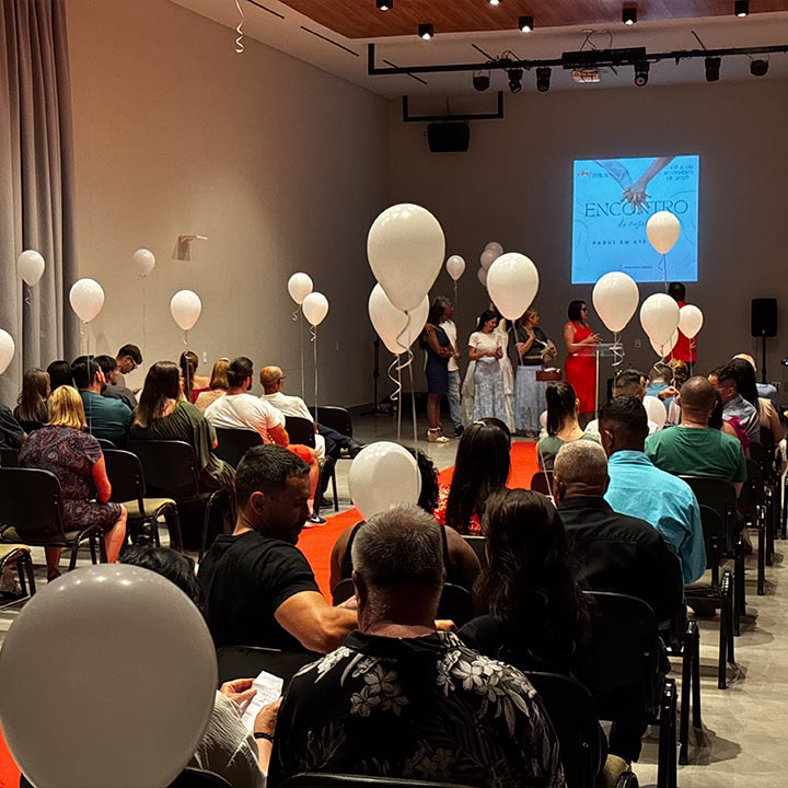 Evento social com pessoas assistindo apresentação, decorado com balões brancos e projeção na parede, mostrando uma reunião ou encontro em um espaço interno moderno.