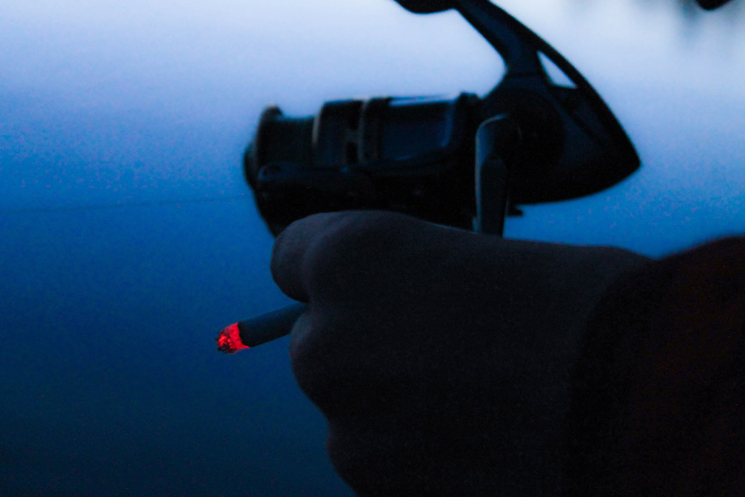 Man holding fishing reel closeup at dusk with lit cigarette between his fingers.