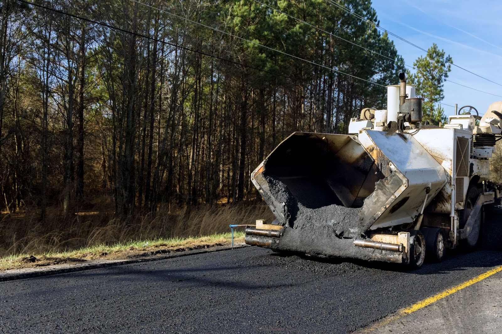 An industrial machinery laying fresh asphalt on the road, with trees lining the side, illustrating the process of asphalt paving in a natural environment.