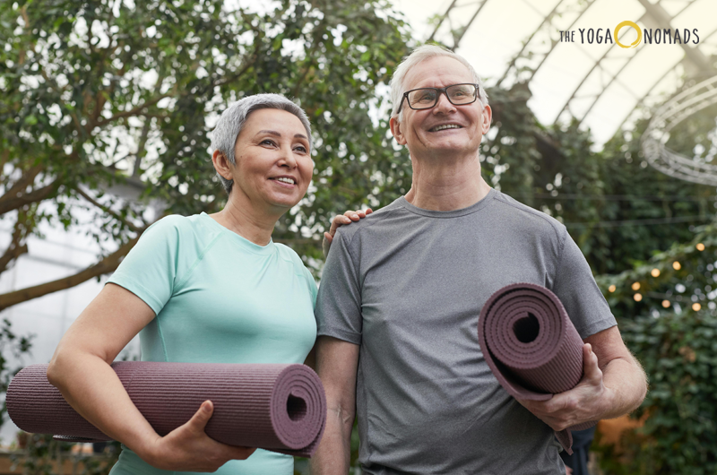 two senior persons standing and smiling while holding their yoga mats