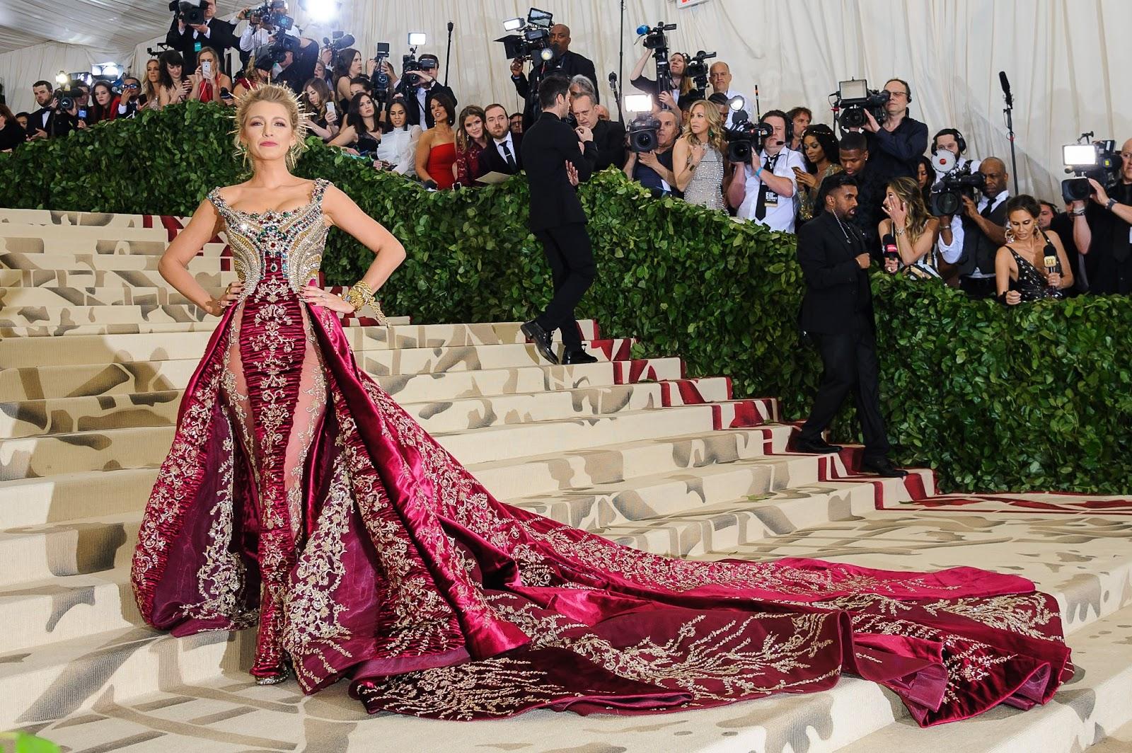 Blake Lively at the 2018 Met Gala wearing a regal burgundy and gold gown with intricate embroidery and a long, dramatic train on the grand staircase.