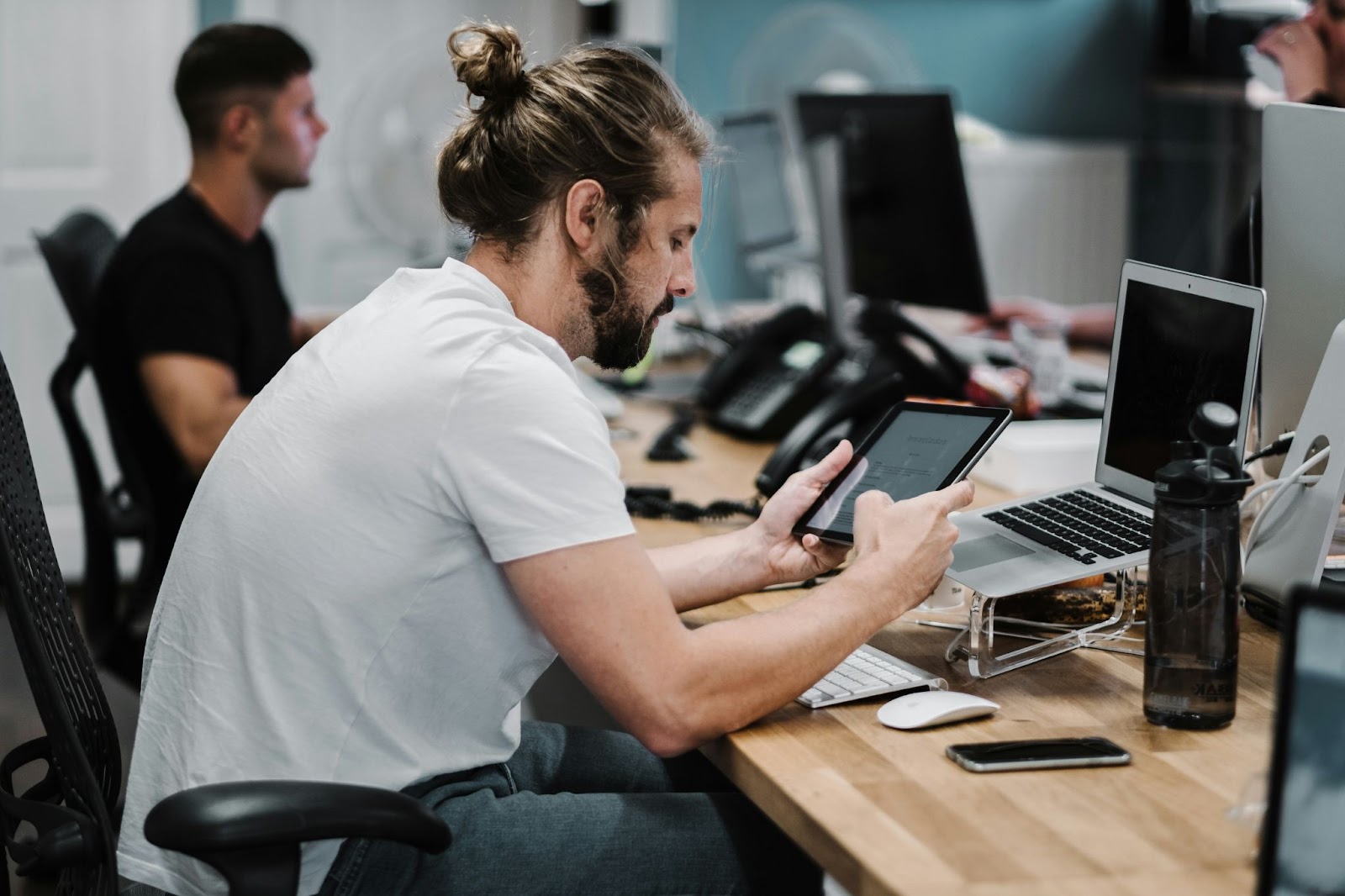 A man is working at a desk with a tablet, laptop, smartphone, and water bottle, while another person works at a computer in the background.