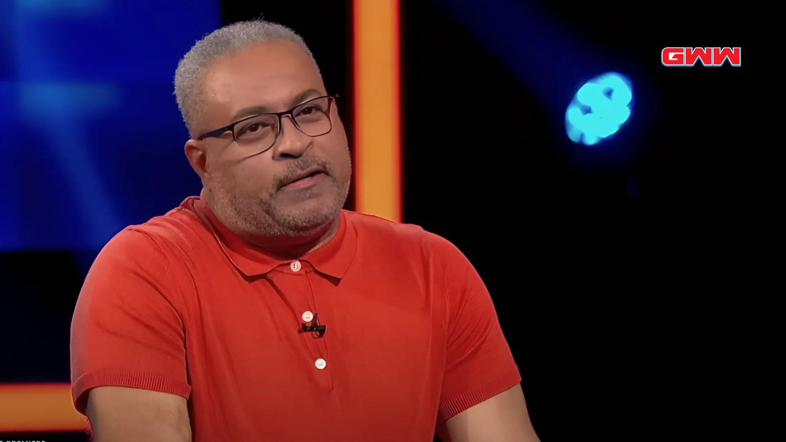 Man wearing red shirt, speaking in front of a dark background.