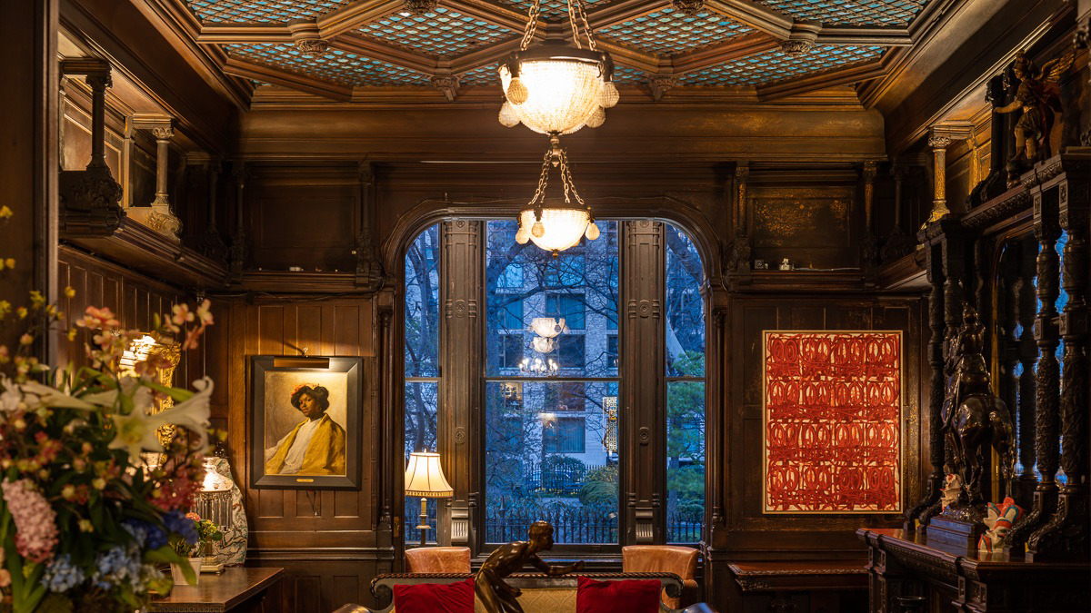 Luxurious, ornately decorated library room at the National Arts Club in New York City, featuring dark wood paneling, a coffered ceiling, elegant chandeliers, large arched windows with city views, classic and modern artwork, and plush seating, creating a sophisticated and historic ambiance.
