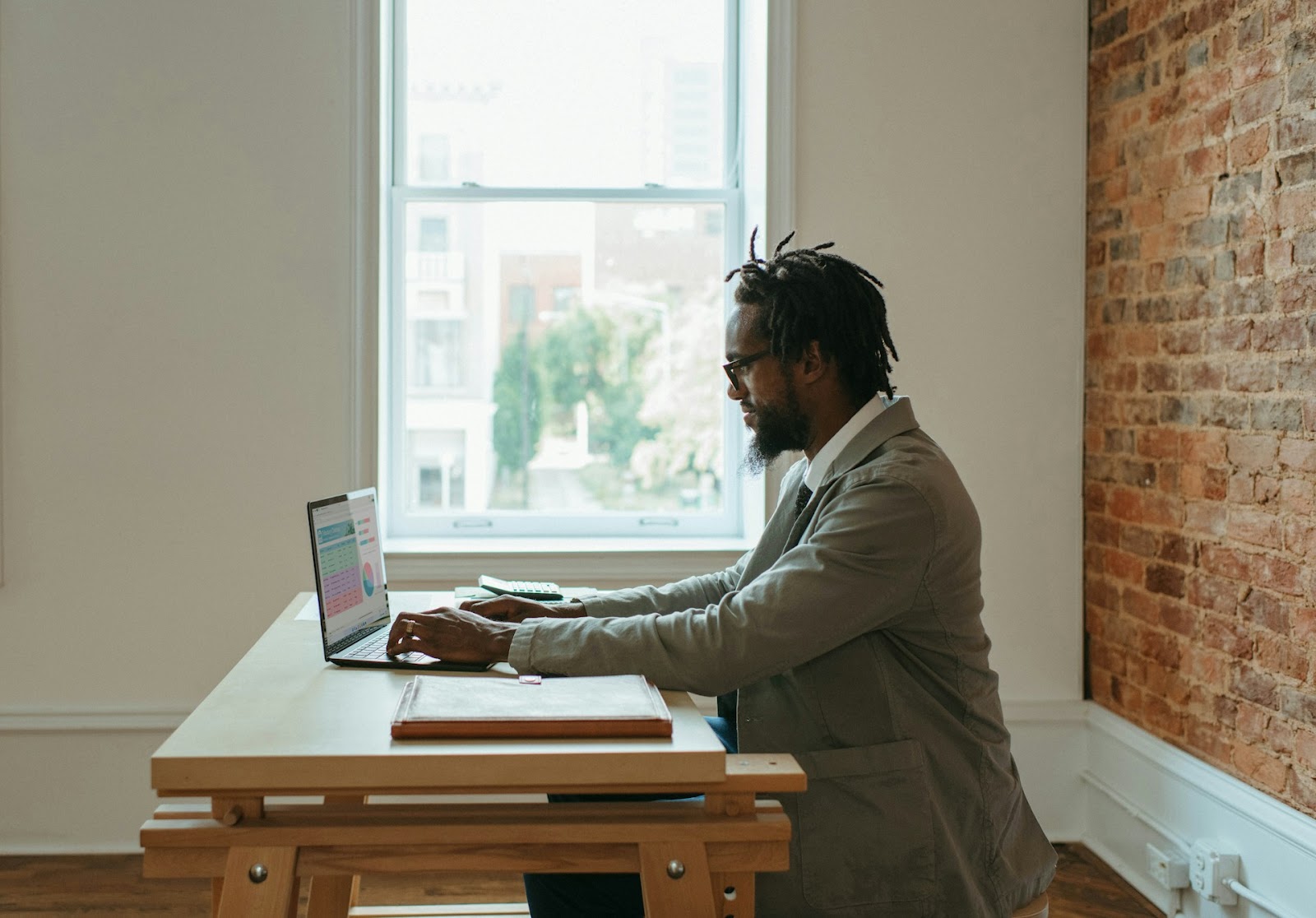 A man focused on his laptop, sitting by a window with natural light streaming in.