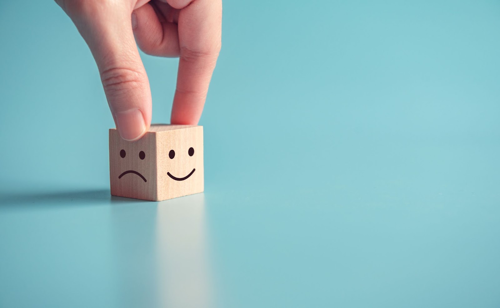 (A hand flipping a wooden block showing a sad face on one side and a happy face on the other, capturing the shift in mood against a light blue background.)