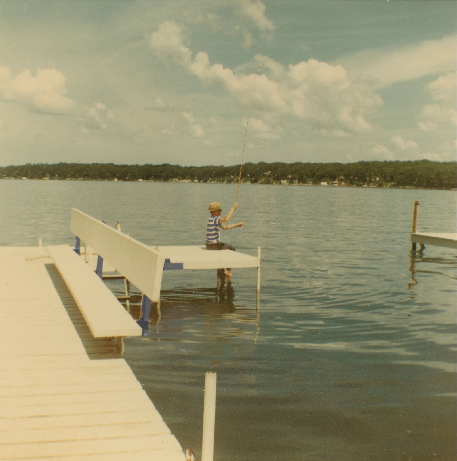 Little boy wearing a blue and white striped shirt sits at the end of a dock, fishing. 