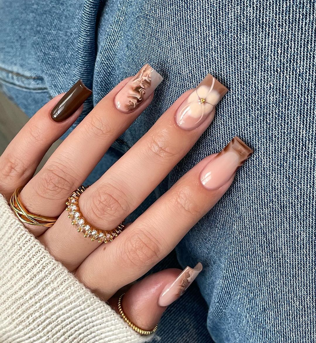 Close-up of a hand with long, decorated nails resting on denim fabric, featuring neutral tones and gold jewelry.

