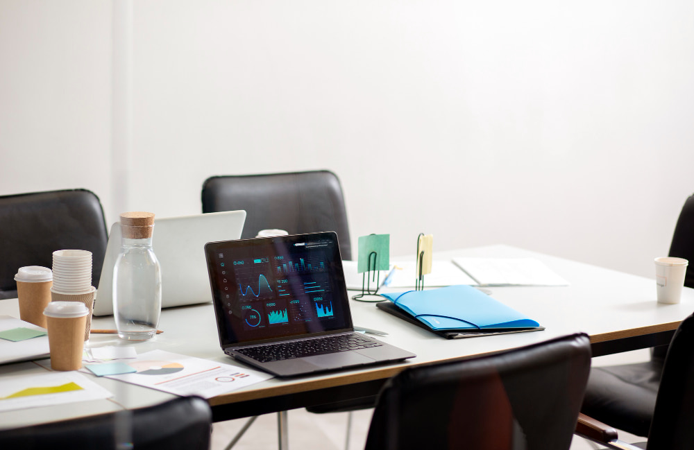 Mesa de reunião com laptops, gráficos em uma tela, pastas, copos de papel e garrafa de vidro, representando um ambiente corporativo 