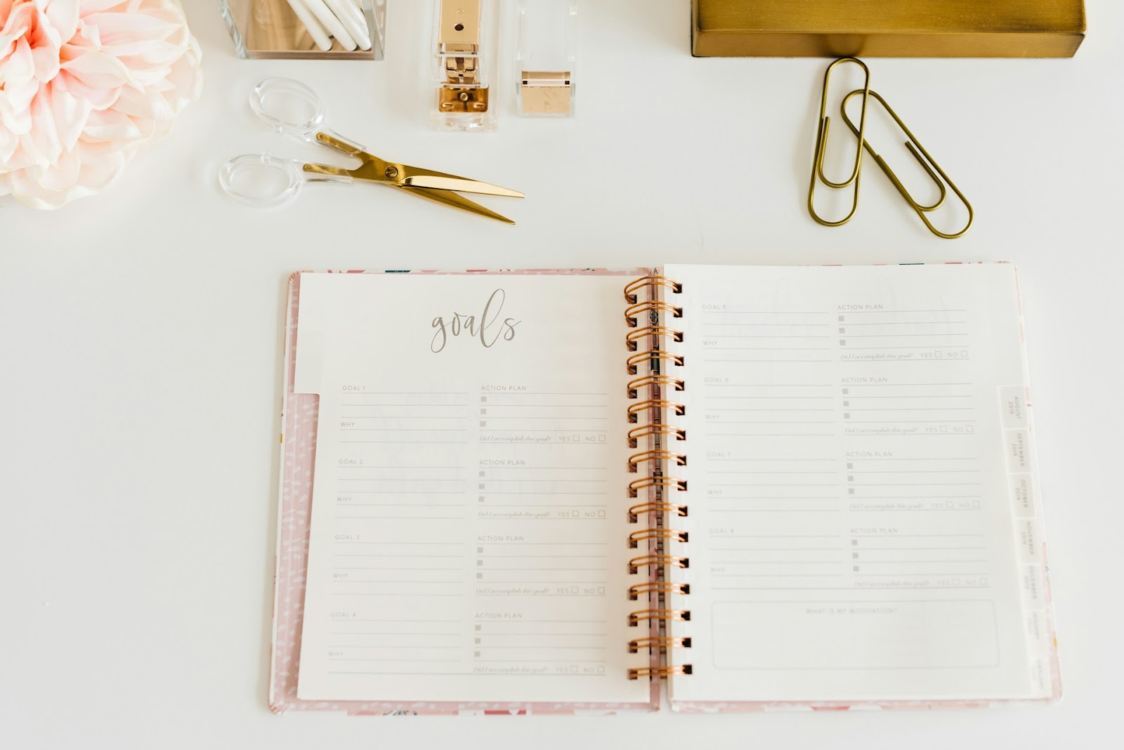 A spiral-bound planner lies open on a desk, displaying a goals section with space for action plans and progress tracking, surrounded by office supplies like scissors, paperclips, and a stapler.