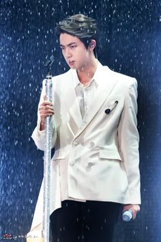 This containsan image of BTS Jin  in a white jacket and black pants holding a microphone while standing under a rain shower