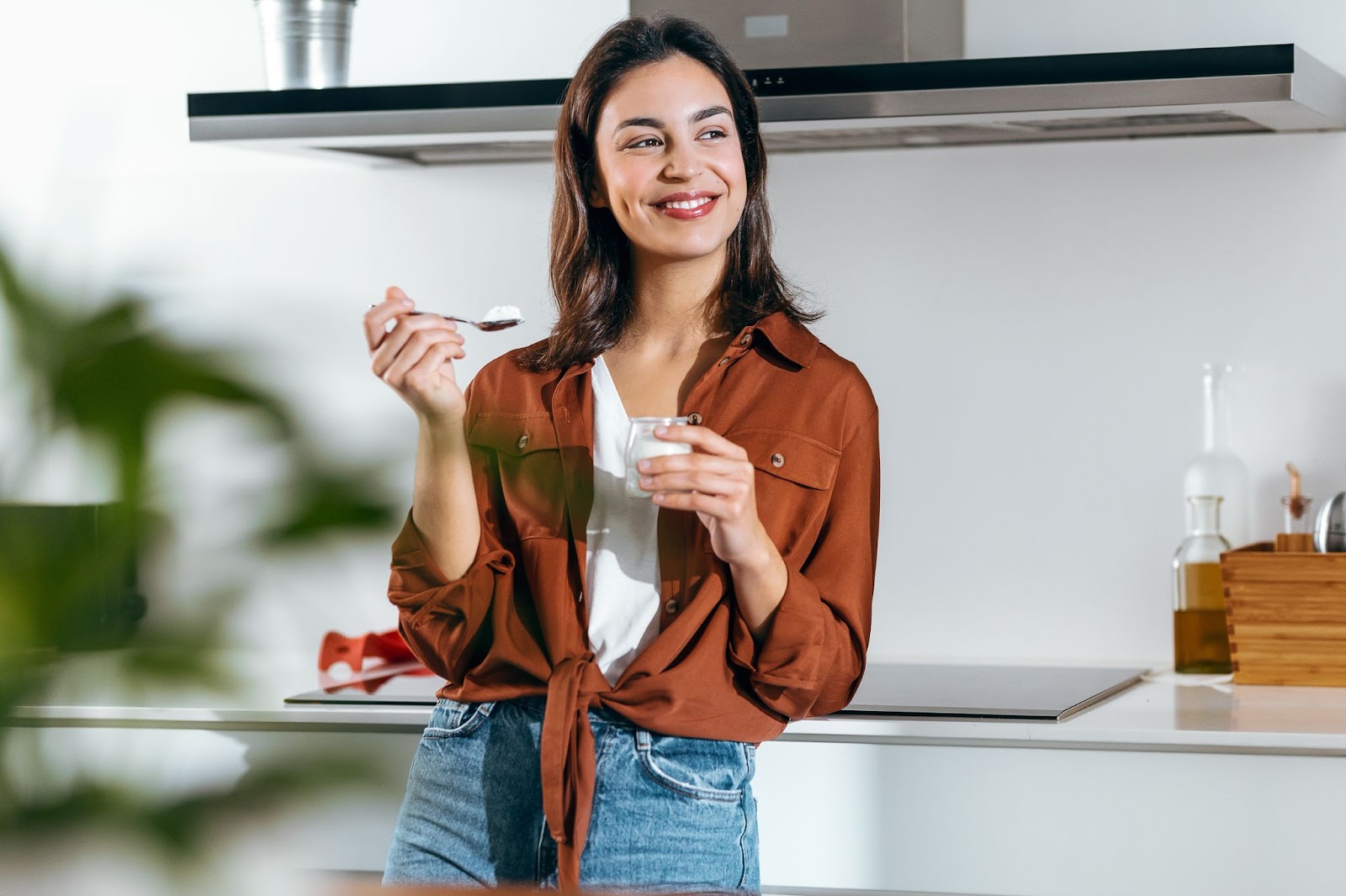 A young woman leaning against her kitchen counter and smiling while eating a cup of yogurt.