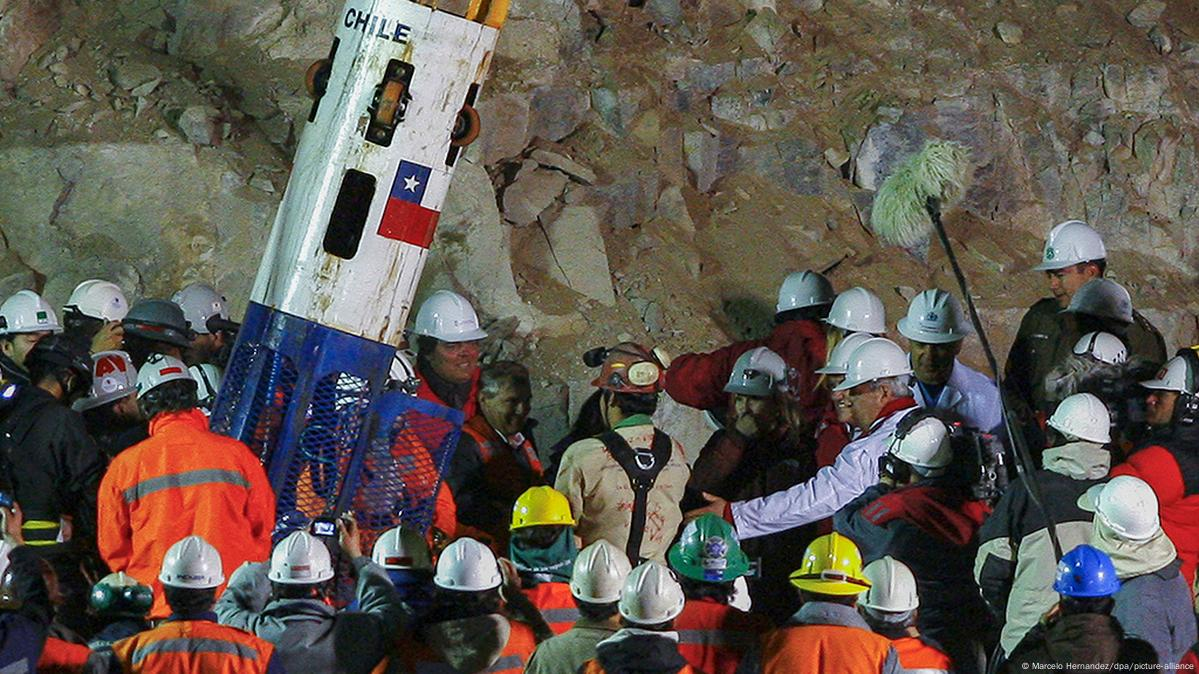 Personas celebrando el rescate de un minero en una cápsula de rescate con bandera de Chile, rodeadas de rocas y trabajadores.