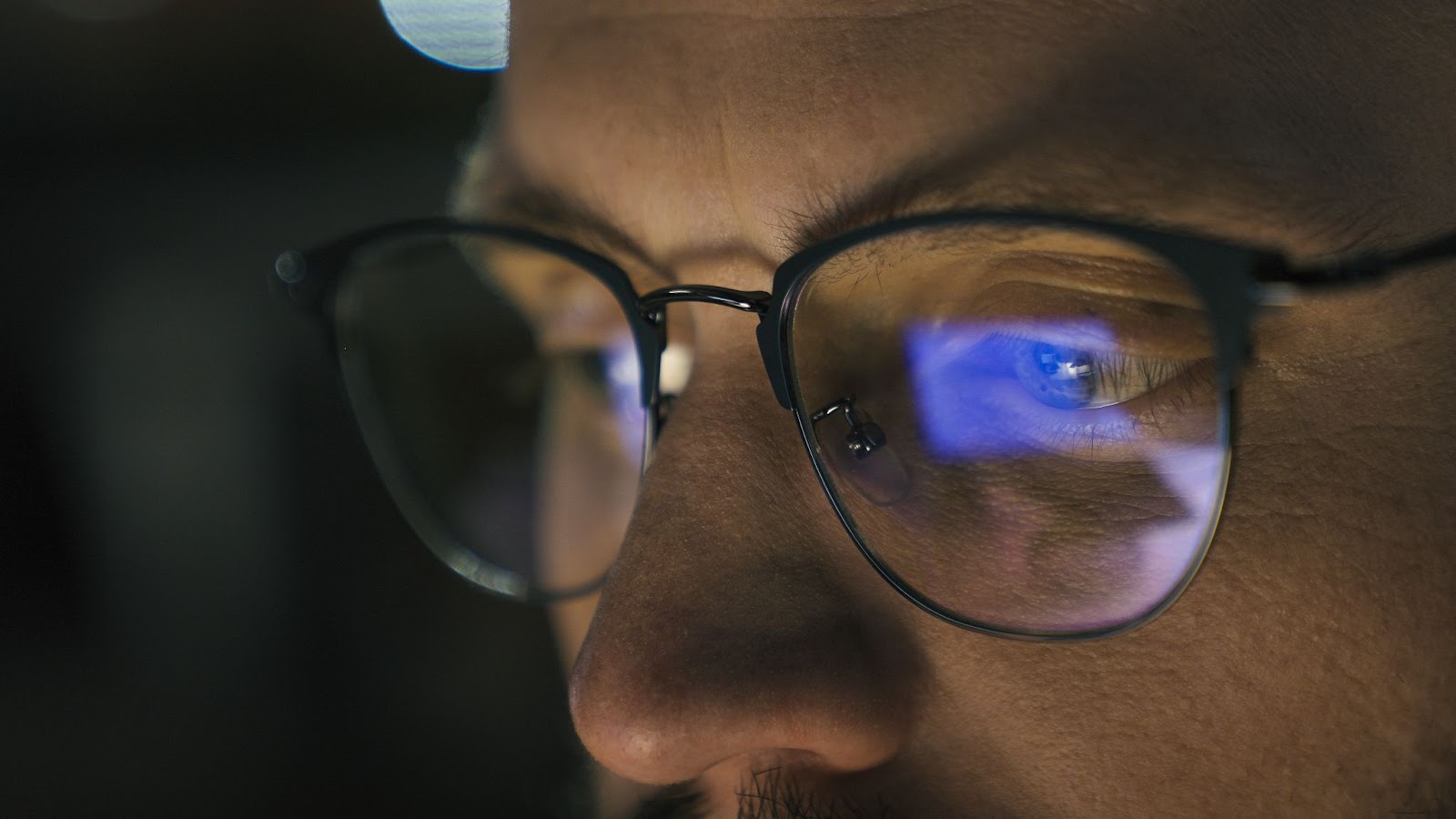 A man's glasses reflect his computer screen as he works in the dark and experiences digital eye strain.