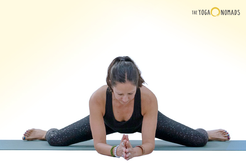 An individual is seated on a yoga mat in a wide-legged forward fold pose, with legs spread wide and torso folded forward, hands clasped together. The background transitions from white to pale yellow