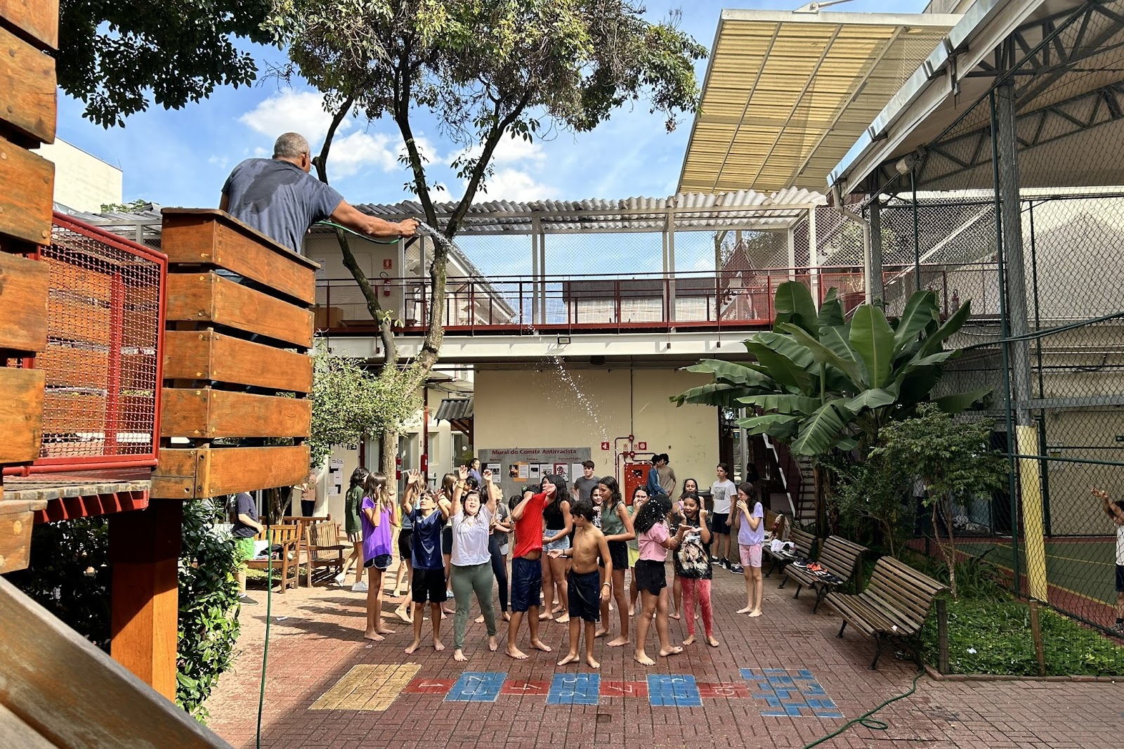 A imagem mostra o pátio de uma escola com crianças tomando banho de mangueira.