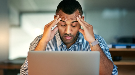 Un homme stressé, assis devant son ordinateur, les mains sur les tempes, manifestant des signes de fatigue et de tension.